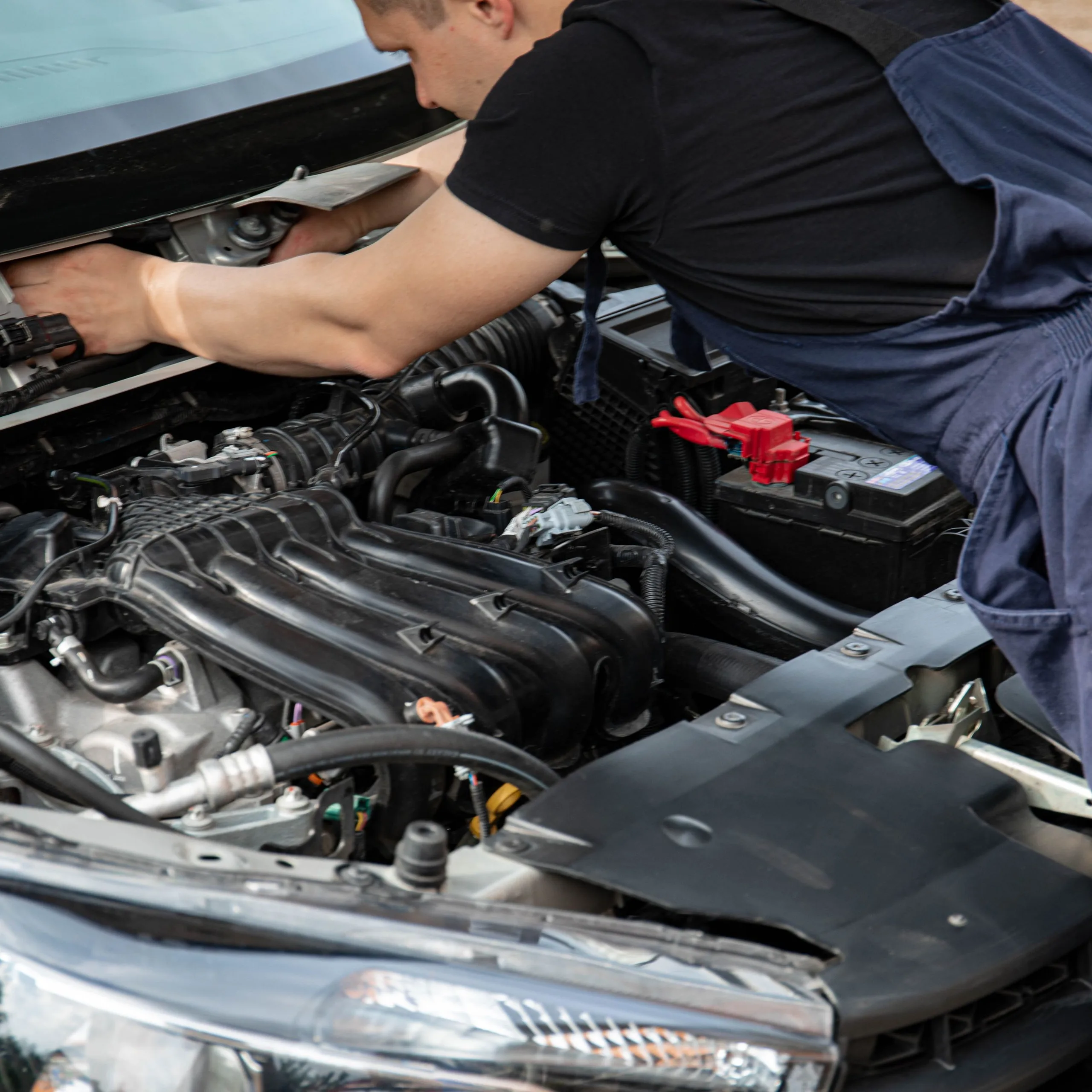 Mécanicien penché sur un capot ouvert : moteur, batterie et phare visibles